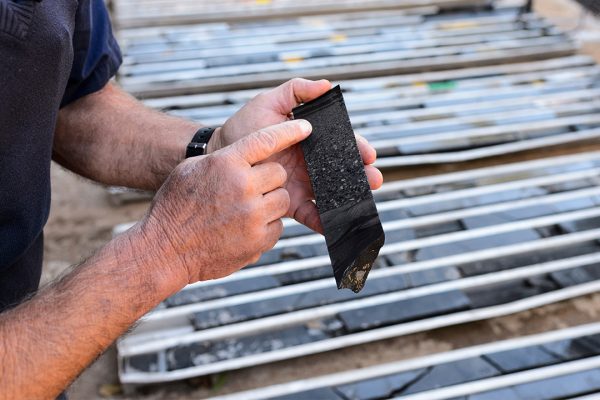 Nic Beukes's hands point to a dark black core sample, trays of samples are out of focus but visible in the background.